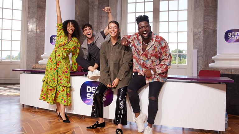 Die vier Jury-Mitglieder der 15. Staffel Dein Song stehen an einem Pult mit Dein Song-Logo angelehnt und blicken fröhlich und lachend in die Kamera.