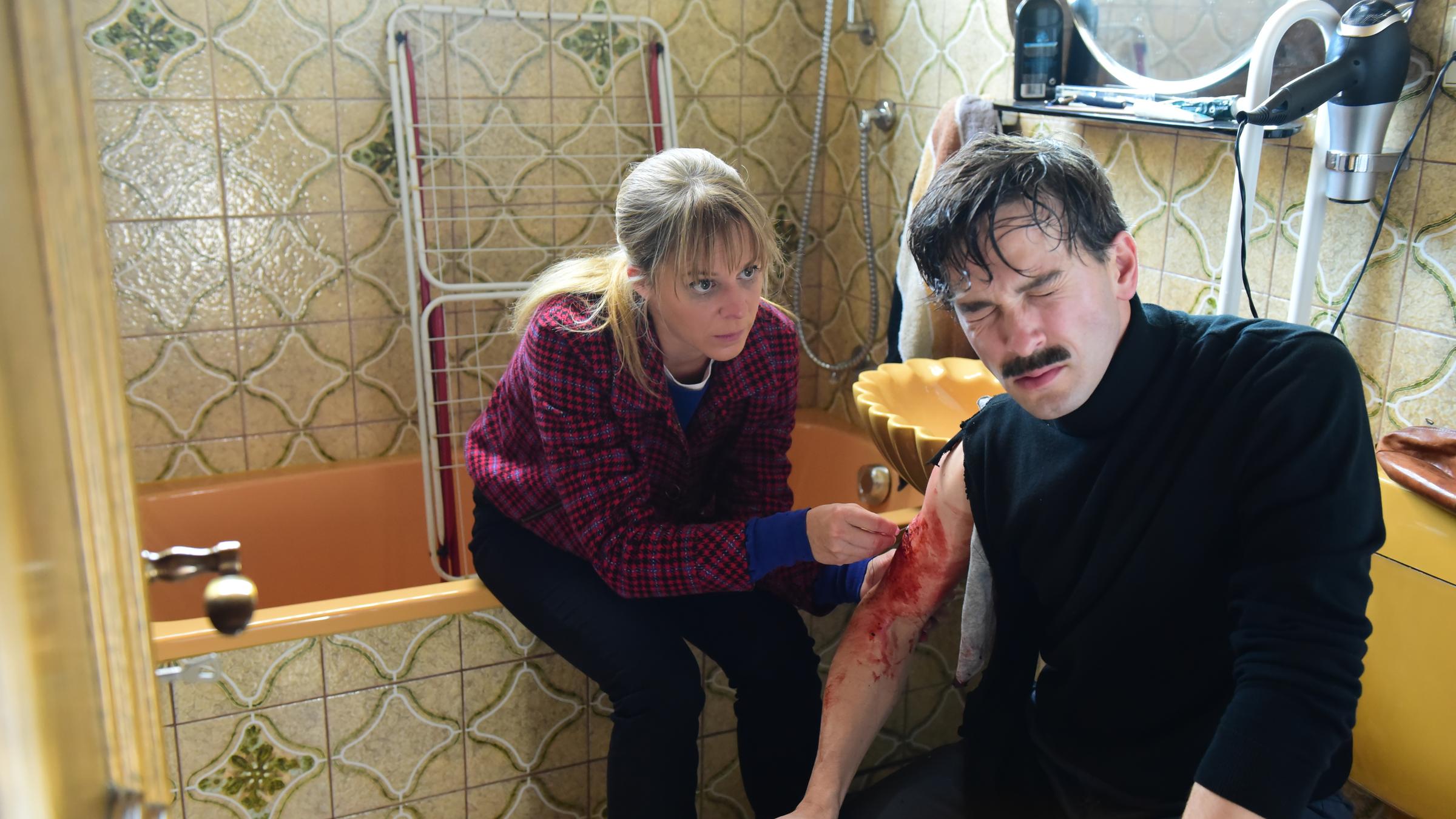 "München Mord – Damit ihr nachts ruhig schlafen könnt": Angelika Flierl (Bernadette Heerwagen) sitzt in einem Badezimmer auf einer Badewanne und näht Mollys (Franz Dinda) blutige Wunde am rechten Arm.