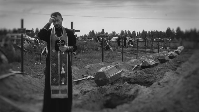 "Das Massaker von Butscha": Schwarz-Weiß-Bild: Im Vordergrund ist ein Priester zu sehen. Hinter ihm stehen mehrere Särge zum Bestatten bereit.