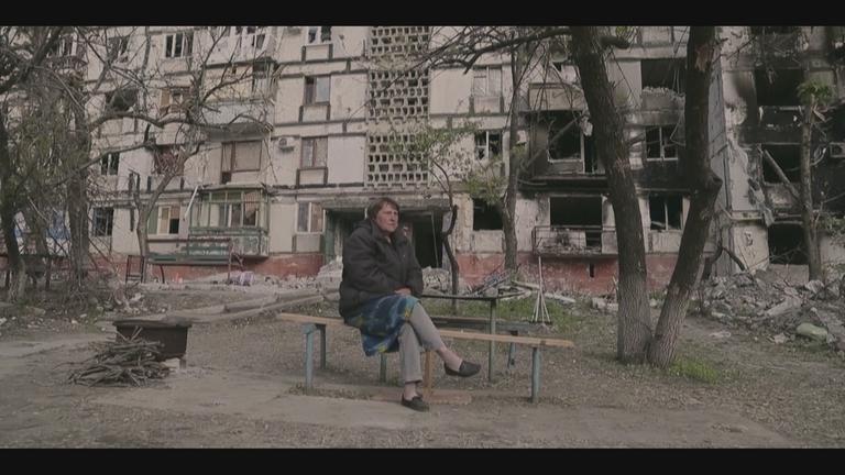 "Krieg gegen die Ukraine – Chronik eines Konfliktes": Eine Frau sitzt auf einer Bank vor einem ausgebrannten Plattenbau.