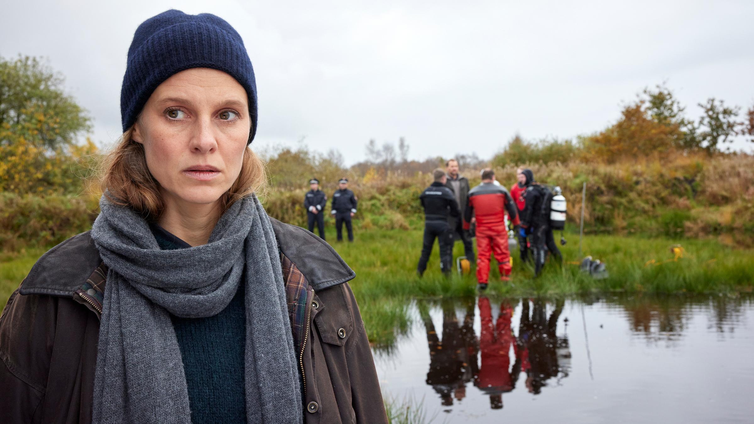 "Ostfriesenmoor": Ann Kathrin Klaasen (Picco von Groote) steht nah vor der Kamera am Moor. Im Hintergrund steht am Rand des Ufers Frank Weller (Christian Erdmann) vor vier Kollegen der Spurensicherung (Komparsen). Hinter ihnen stehen abseits zwei uniformierte Polizisten (Komparsen).