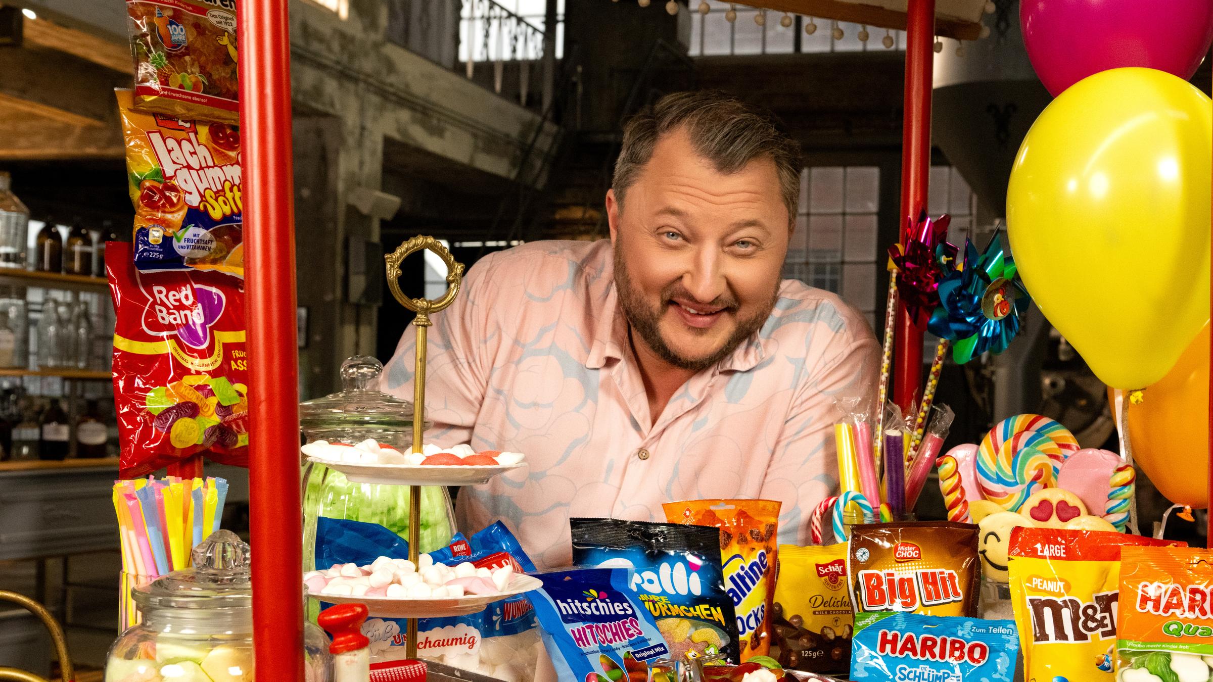 "ZDFzeit: Die Tricks von Haribo, Ferrero & Co. - ZDFbesseresser: Sebastian Lege deckt auf": Sebastian Lege steht hinter einem bunten Stand. In der Auslage befinden sich eine Menge verschiedenster Süßigkeiten.