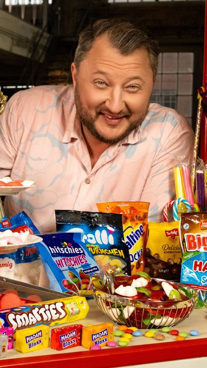 "ZDFzeit: Die Tricks von Haribo, Ferrero & Co. - ZDFbesseresser: Sebastian Lege deckt auf": Sebastian Lege steht hinter einem bunten Stand. In der Auslage befinden sich eine Menge verschiedenster Süßigkeiten.