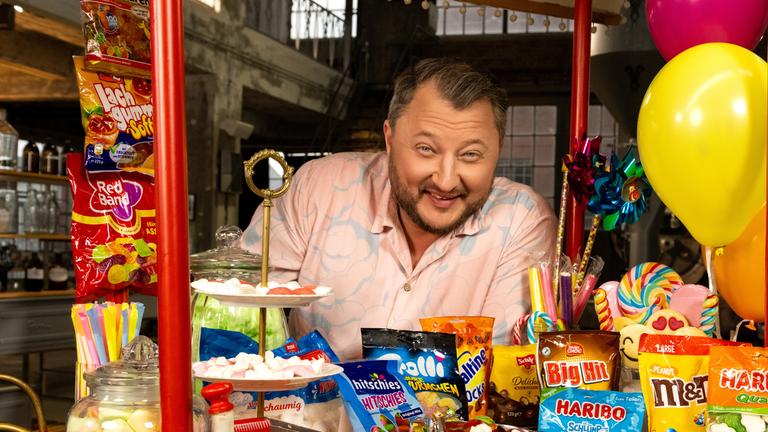 "ZDFzeit: Die Tricks von Haribo, Ferrero & Co. - ZDFbesseresser: Sebastian Lege deckt auf": Sebastian Lege steht hinter einem bunten Stand. In der Auslage befinden sich eine Menge verschiedenster Süßigkeiten.
