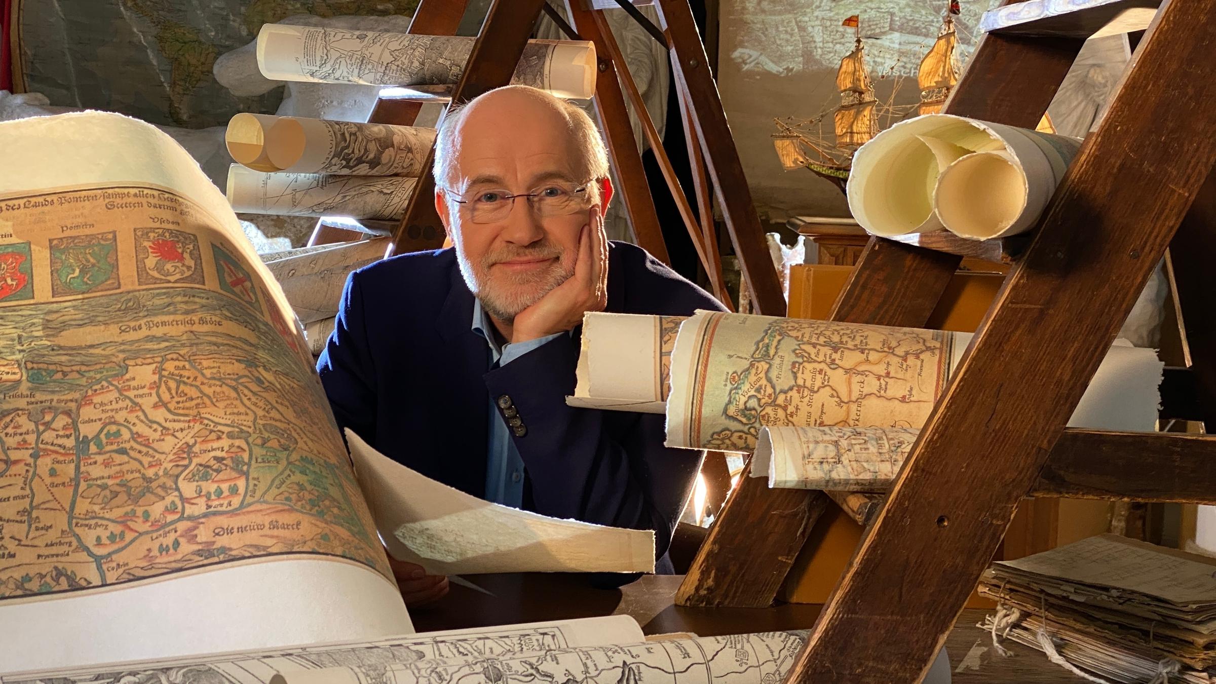 "Ungelöste Fälle der Archäologie - Verschollen": Moderator Harald Lesch sitzt in einem Raum, umgeben von Landkarten, und lächelt in die Kamera.