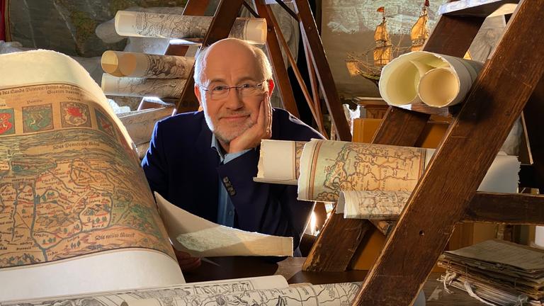 "Ungelöste Fälle der Archäologie - Verschollen": Moderator Harald Lesch sitzt in einem Raum, umgeben von Landkarten, und lächelt in die Kamera.