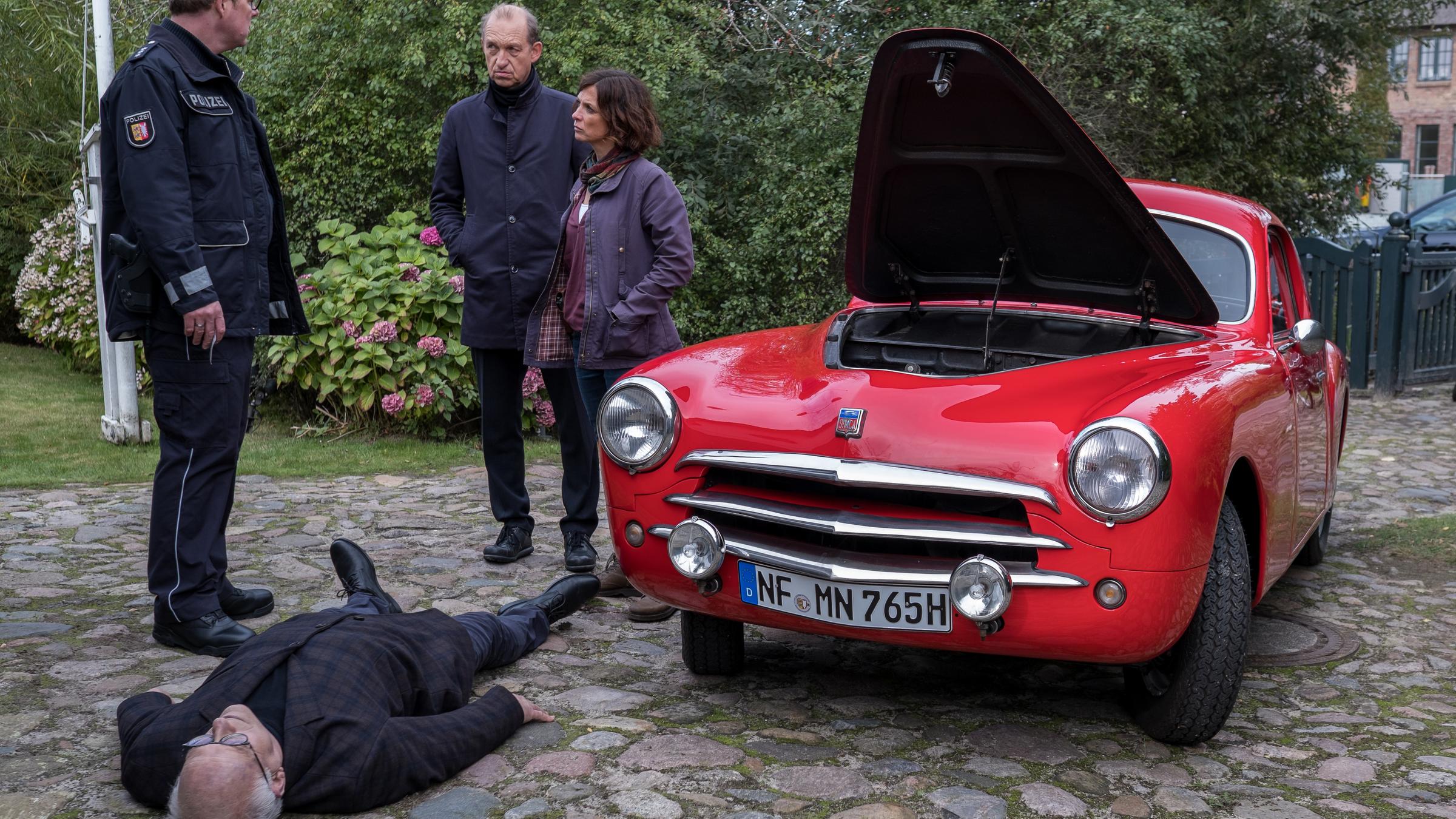 "Nord Nord Mord - Sievers und der große Knall": Carl Sievers (Peter Heinrich Brix), Ina Behrendsen (Julia Brendler) und Kollege Schneider (Stephan A. Tölle) stehen auf einer gepflasterten Hauseinfahrt neben einem schicken knallroten Oldtimer. Die Motorhaube ist noch geöffnet. Zwischen Ihnen liegt der tote Friedel Tanner (Dieter Flechsenberger) ausgestreckt auf dem Boden.