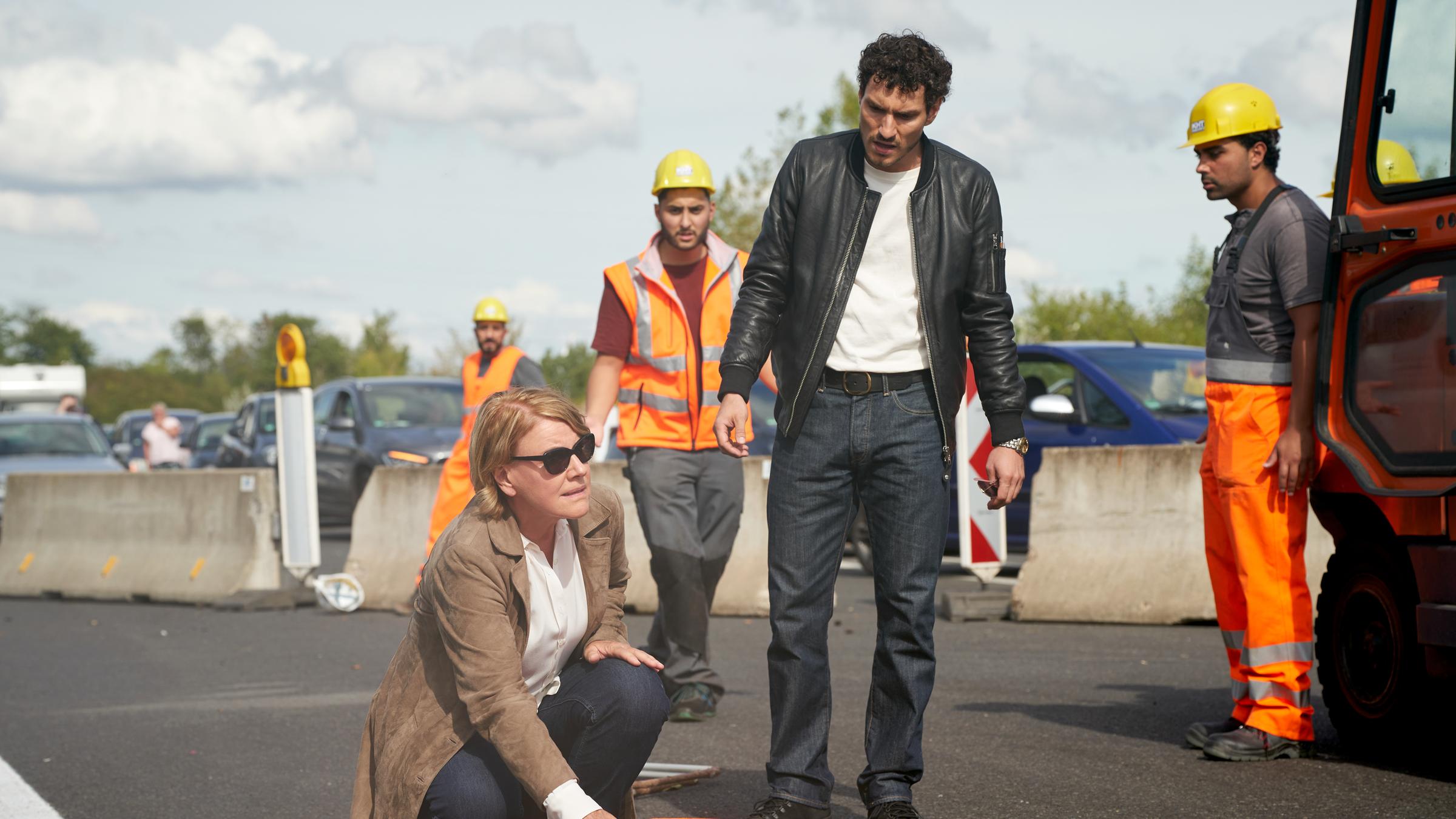 "Marie Brand und die Ehrenfrauen": Marie Brand (Mariele Millowitsch) kniet vor der dem Verletzten Vorarbeiter Claudio Messina (Sami Loris) und prüft mit ihrer rechten Hand ob er noch einen Puls hat. Giuseppe Costa (Nicola Perot, rechts) steht daneben. Im Hintergrund sieht man drei Bauarbeiter (Komparsen).
