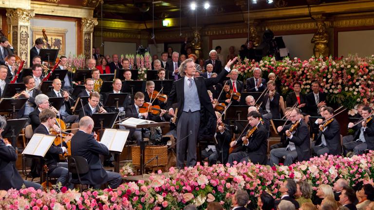 "Neujahrskonzert der Wiener Philharmoniker 2023": Dirigent Franz Welser-Möst steht im Frack dem Publikum zugewandt mit dem Rücken zum Orchester. Er hält in der rechten Hand den Dirigierstab, den linken Arm streckt er in die Höhe, um das Publikum zum Klatschen anzuregen.