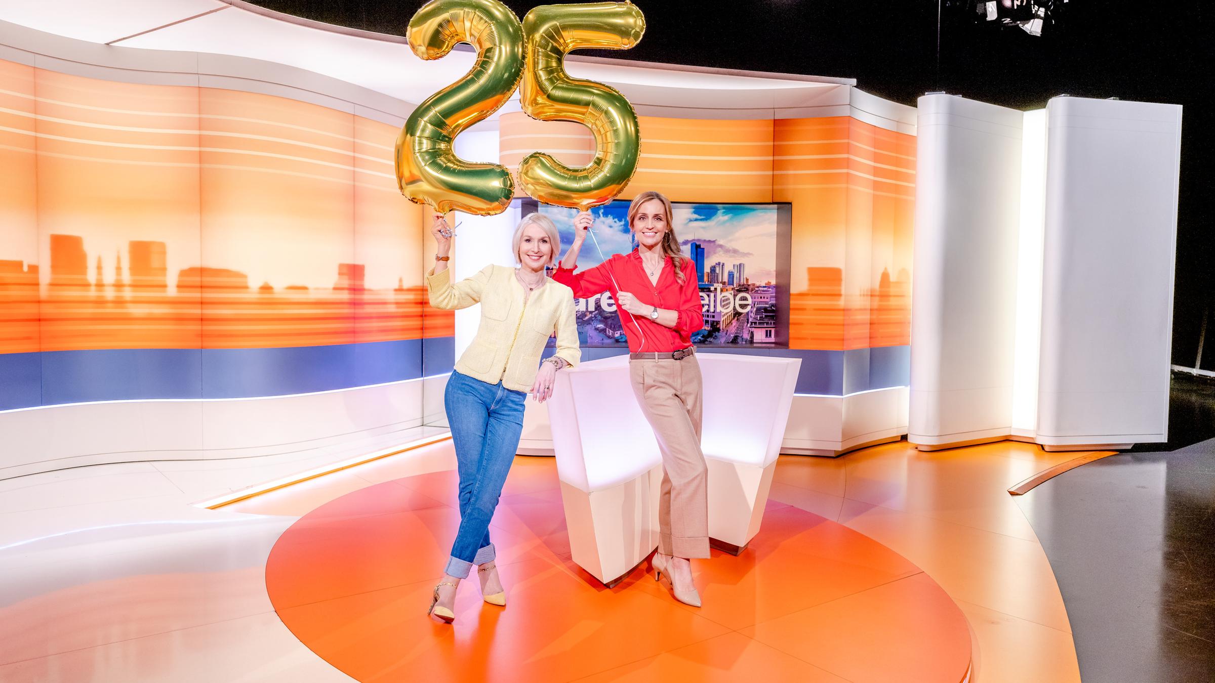 25 Jahre drehscheibe: Babette von Kienlin und Sandra Maria Gronewald im Studio. Sie halten zwei goldene Luftballons in die Höhe, die die Zahl "25" bilden.