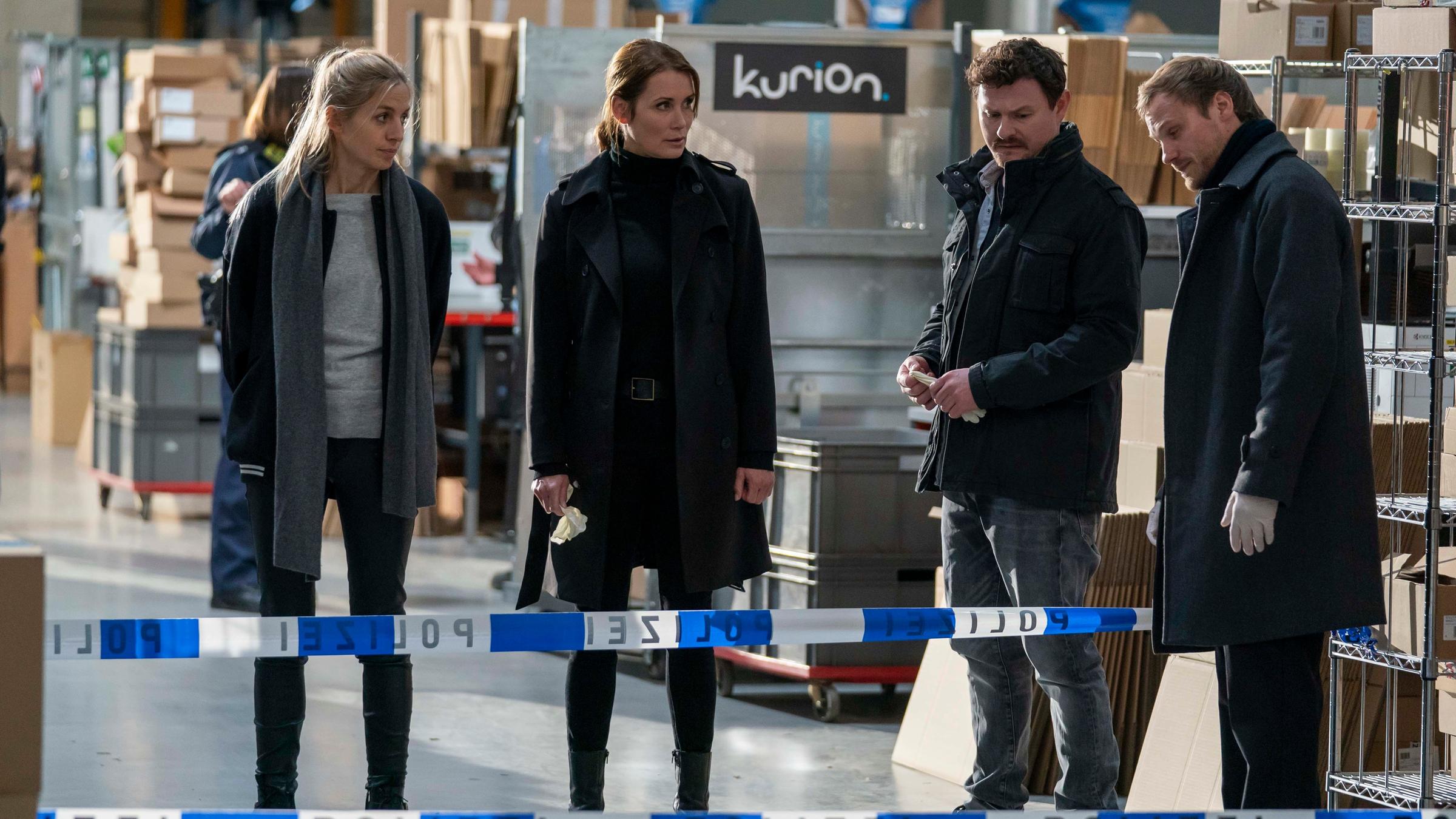 "Das Quartett - Tödliche Lieferung": Pia Walther (Annika Blendl), Maike Riem (Anja Kling), Christoph Hofherr (Shenja Lacher) und Linus Roth (Anton Spieker) stehen in einer Lagerhalle und blicken auf die auf dem Boden liegende Leiche eines Mannes. In den riesigen Regalen im Hintergrund stapeln sich Pakete.