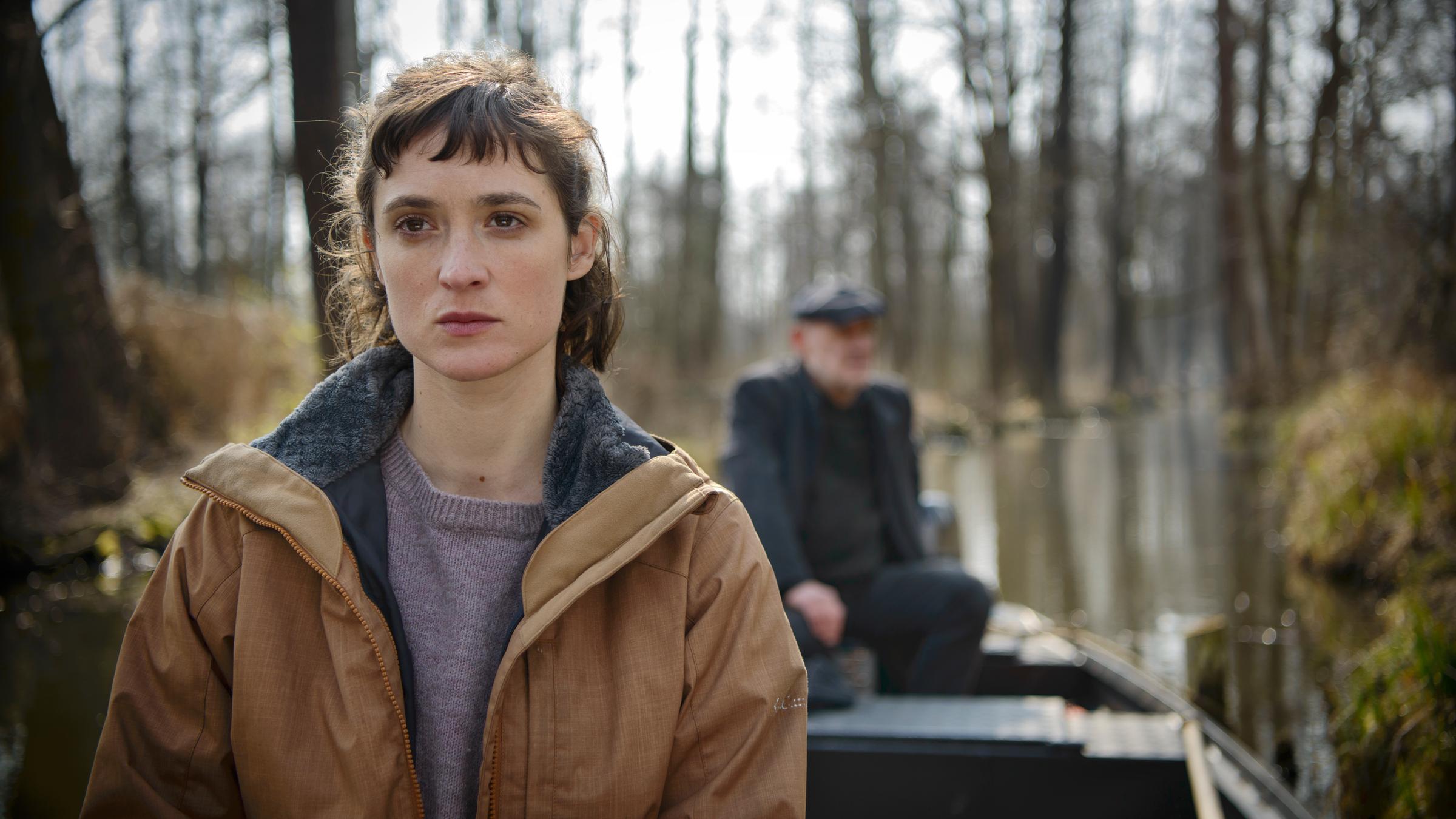 "Spreewaldkrimi - Die 7. Person": Maja Wichmann (Friederike Becht) sitzt in einem Spreewaldkahn und blickt skeptisch in die Kamera. Im Hintergrund sitzt Kommissar Krüger (Christian Redl) an der Pinne des Außenbordmotors und steuert den Kahn durch den Fließ.