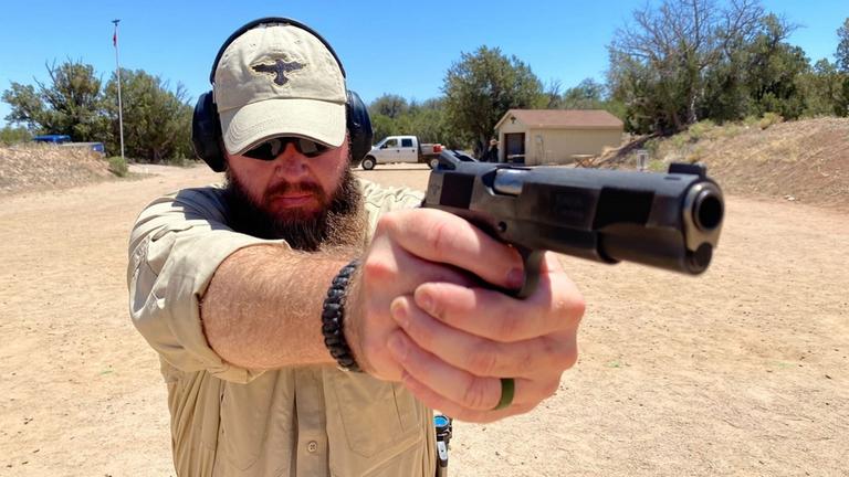 "Gun Nation - Amerikas tödlicher Waffenwahn": Ein bärtiger Mann in sandfarbener Kleidung und mit Schutzbrille und Kopfhörern zielt mit einer Feuerwaffe rechts an der Kamera vorbei.