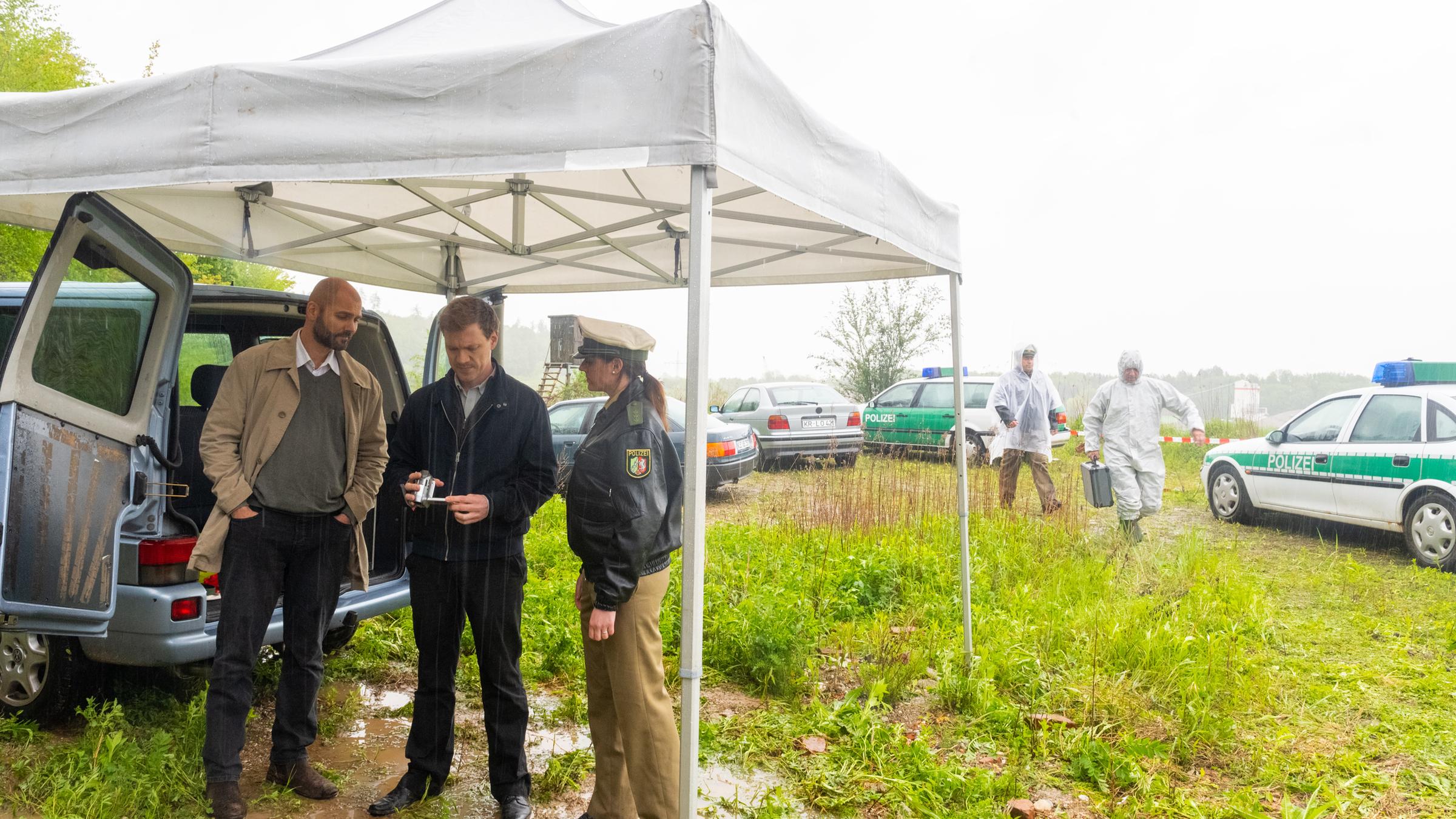 "XY gelöst - Mörderische Konflikte": Nachgestellte Szene. Ein regnerischer Tag auf einer abgesperrten Wiese. Zwei Ermittler stehen unter einem Pavillon. Neben ihnen steht eine Polizistin. Alles drei schauen konzentriert auf das Handy einer der Ermittler. Vor der Absperrung stehen zwei Autos und zwei Polizeiwagen.