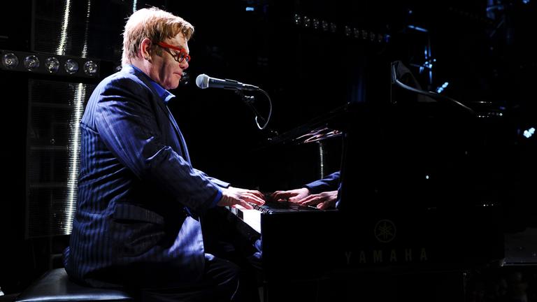 "The True Story of Elton John": Elton John trägt eine rote Sonnenbrille und ein gestreiftes blaues Sakko. Er spielt Klavier.