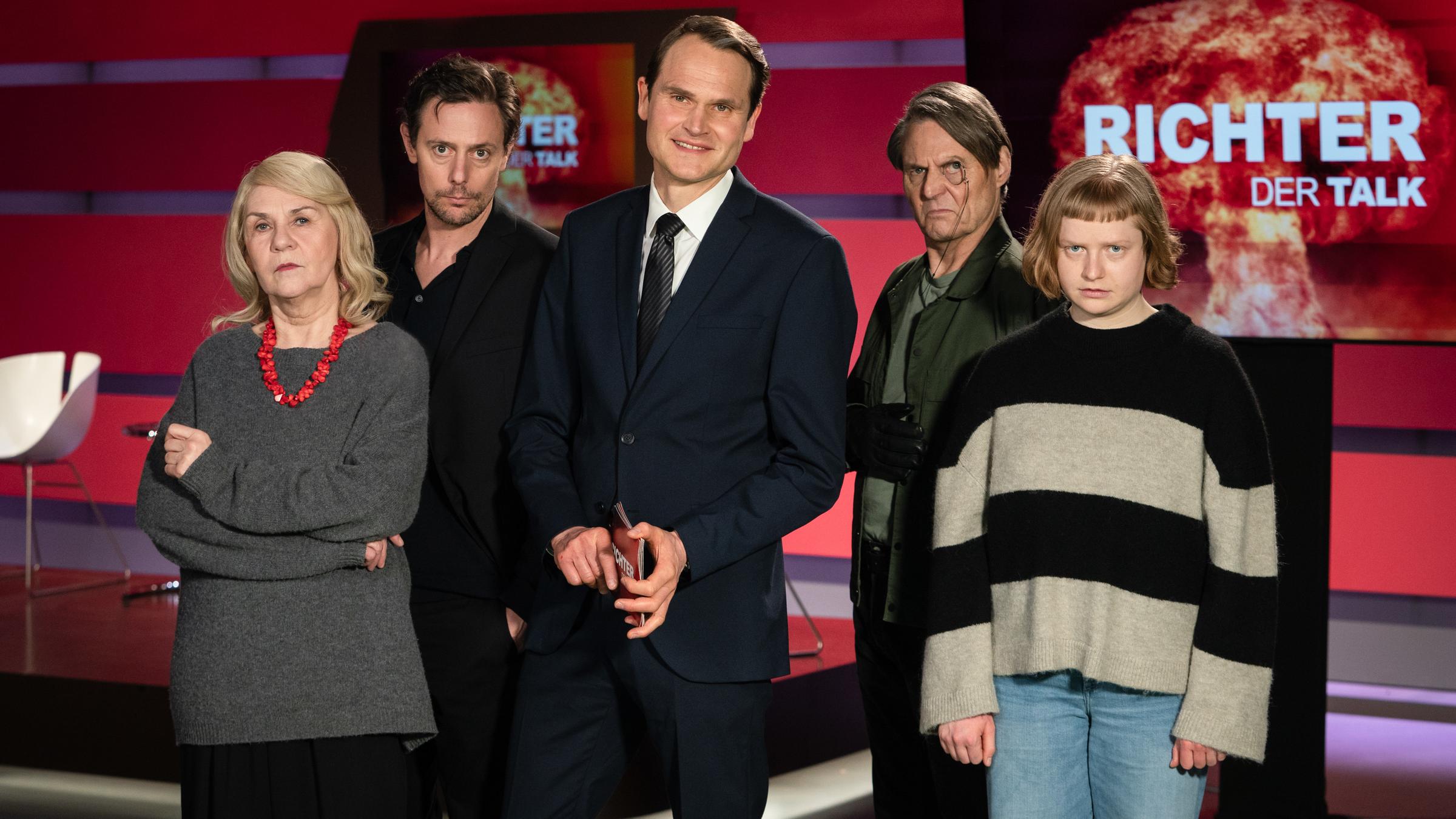 "Browser Ballett: Atomkrieg – Der Talk": Barbara Schmitz (Birgit Berthold), Volker Recht (Roland Bonjour), Magnus Richter (Fabian Hinrichs), Jürgen Hahn (Wolfgang Bahro) und Clara Neumann (Luise von Stein) stehen im Studio von "Richter – Der Talk". Auf den Bildschirmen im Hintergrund ist eine riesige Explosion zu sehen. Während seine Gäste eher streng schauen, lächelt Richter in die Kamera.