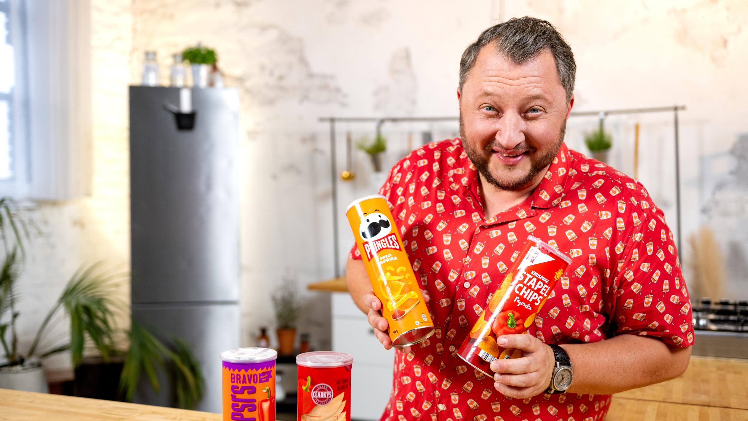 "BesserEsser - Lege packt aus: Sündige Snacks": Sebastian Lege steht in einem Küchenset und grinst in die Kamera. In jeder Hand hält er eine Packung Stapelchips, vor ihm stehen nochmal zwei Packungen.