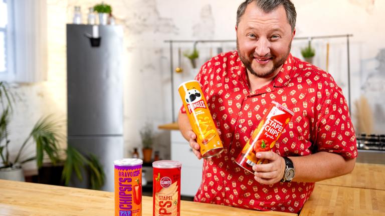 "BesserEsser - Lege packt aus: Sündige Snacks": Sebastian Lege steht in einem Küchenset und grinst in die Kamera. In jeder Hand hält er eine Packung Stapelchips, vor ihm stehen nochmal zwei Packungen.