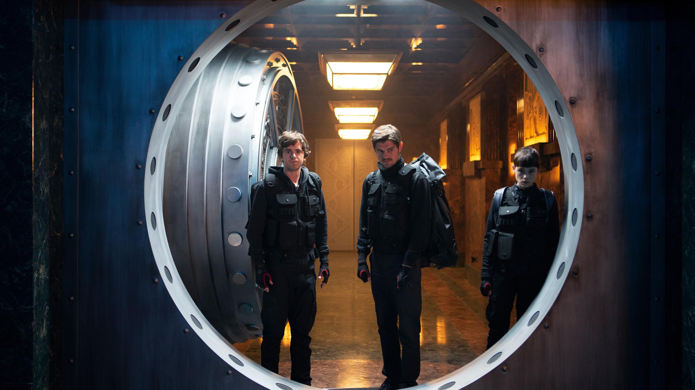  "Countdown in Madrid": Halbtotale. Thom (Freddie Highmore), James (Sam Riley) und Lorraine (Àstrid Bergès-Frisbey) stehen mit angespannten Blicken in offener Tür von Tresorraum. Die Kamera blickt aus dem Tresorraum heraus auf das Trio.