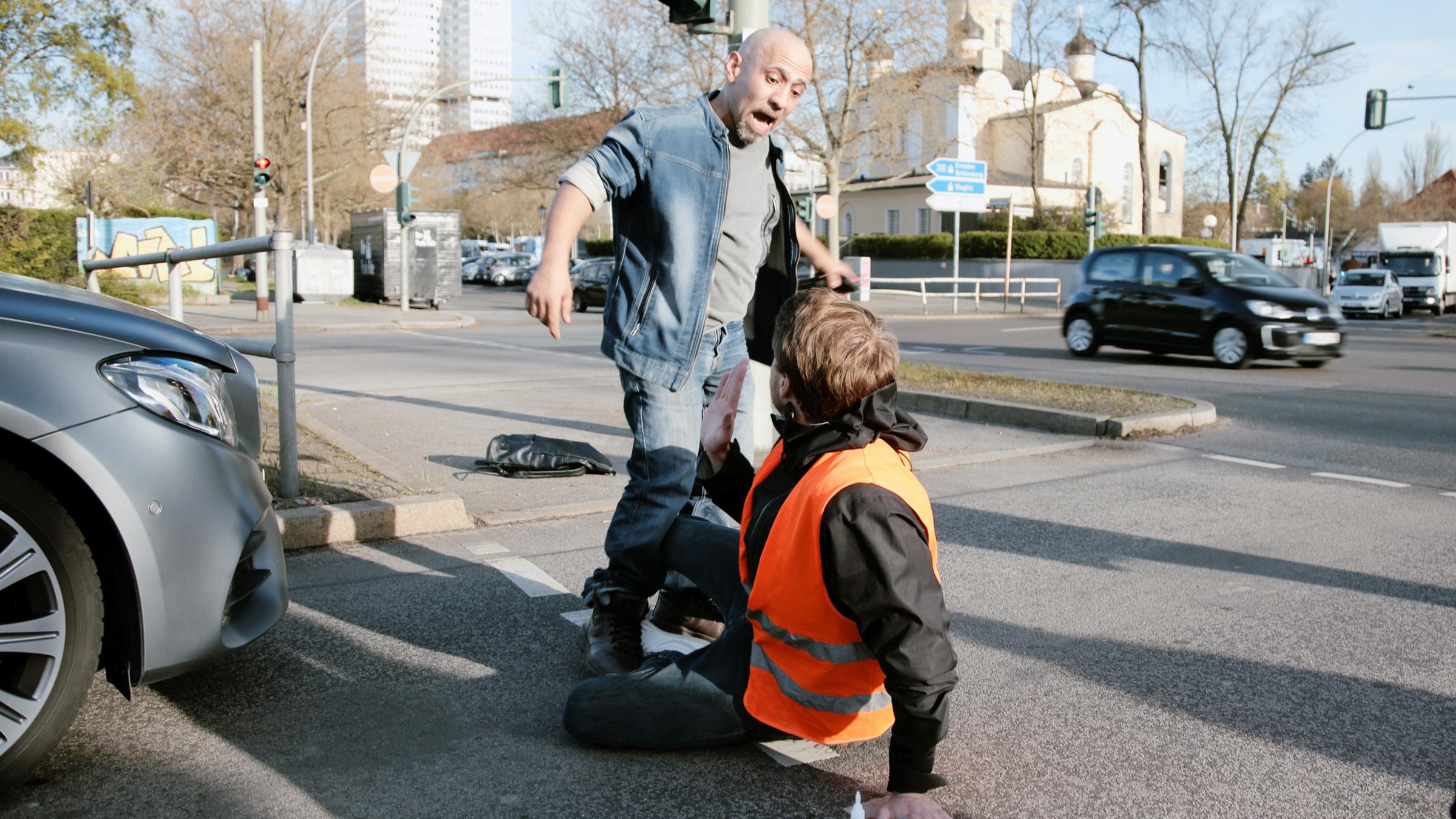 "Radikal, gehasst, verzweifelt- Letzte Generation": Ein junger Mann sitzt auf der Fahrbahn und hat seine Hand festgeklebt. Der stehende Mann neben ihm, ist wütend.