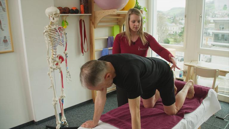 "Heilung für chronisch Kranke? - Vom Profit zur Prävention": Rückenschmerzpatient kniet auf der Physiotherapie-Liege, die Therapeutin leitet ihn an.