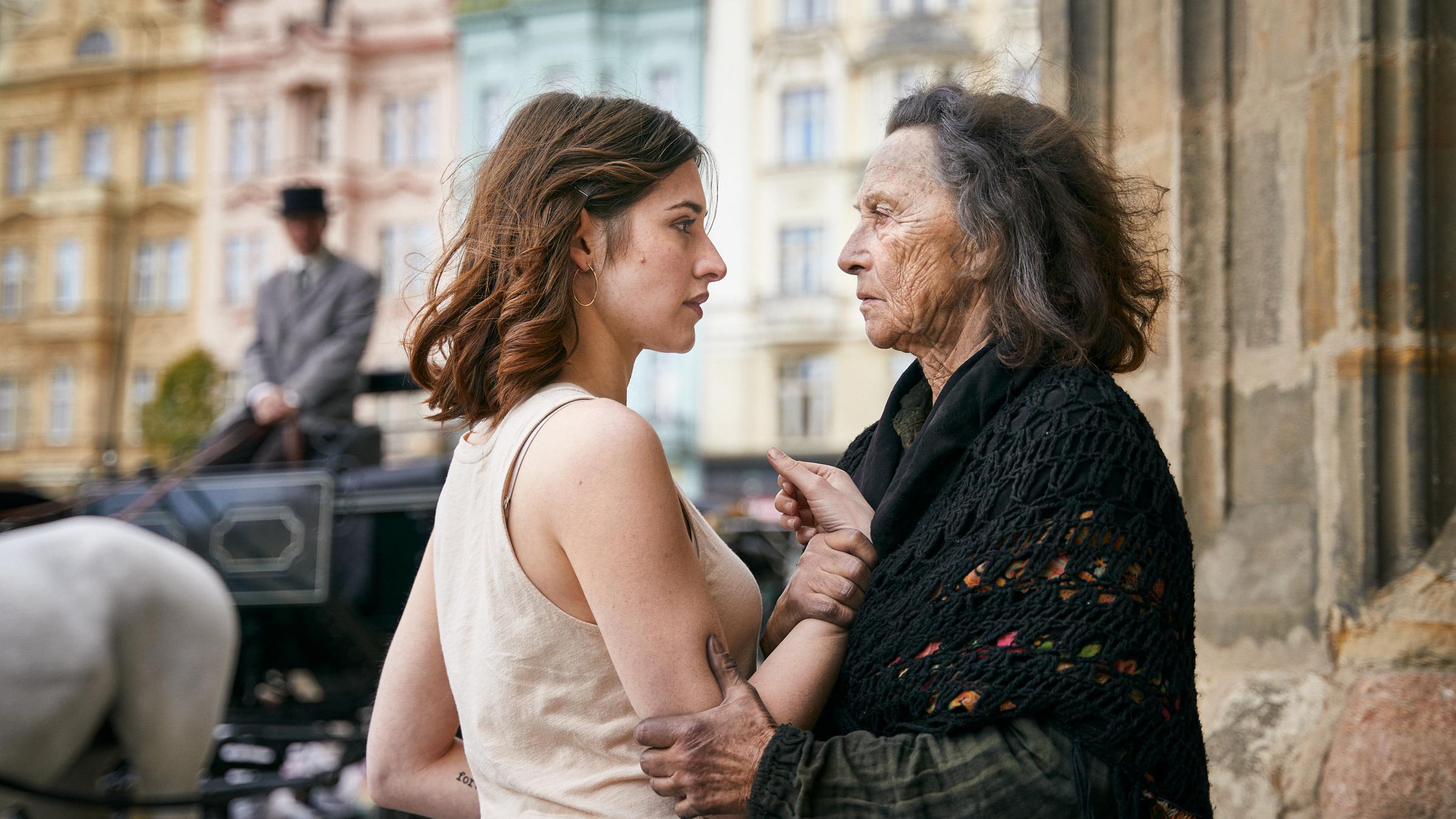 "Der Schatten - Die Prophezeiung": Norah Richter (Deleila Piasko) wird von der Bettlerin Dorothea Lechner (Elisabeth Rath) fest am Arm gepackt. Die beiden stehen sich gegenüber und schauen sich eindringlich in die Augen. Im Hintergrund sieht man eine Kutsche vor einer Hausfassade.