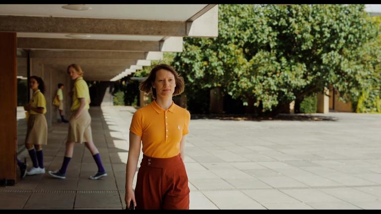 "Club Zero": Miss Novak (Mia Wasikowska) läuft unter der überdachten Balustrade eine Schulgebäudes. Im Hintergrund drei Schülerinnen und Schüler in Schulkeidung.