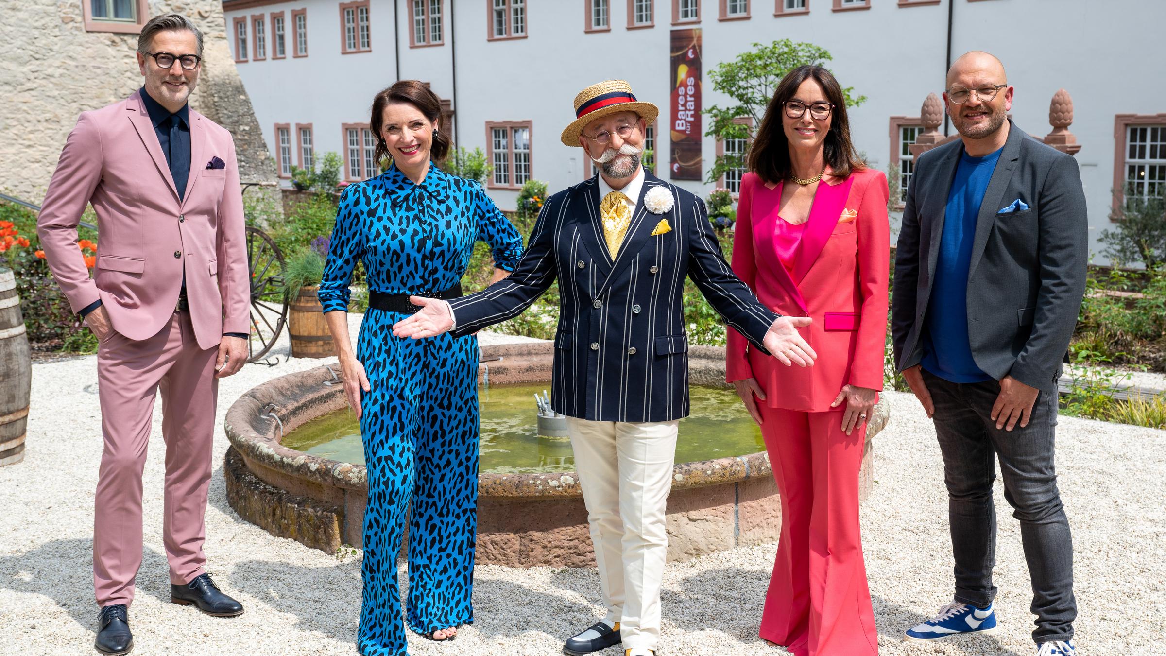 "Bares für Rares - Deutschlands größte Trödelshow": Detlev Kümmel, Wendela Horz, Horst Lichter, Dr. Heide Rezepa-Zabel und Sven Deutschmanek stehen nebeneinander und lächeln in die Kamera. Horst Lichter hat die Arme einladend gen Kamera ausgebreitet. Im Hintergrund ist ein Brunnen, Rosen sowie Gebäude des Kloster Eberbachs zu sehen.