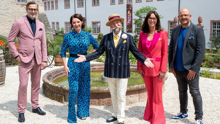 "Bares für Rares - Deutschlands größte Trödelshow": Detlev Kümmel, Wendela Horz, Horst Lichter, Dr. Heide Rezepa-Zabel und Sven Deutschmanek stehen nebeneinander und lächeln in die Kamera. Horst Lichter hat die Arme einladend gen Kamera ausgebreitet. Im Hintergrund ist ein Brunnen, Rosen sowie Gebäude des Kloster Eberbachs zu sehen.