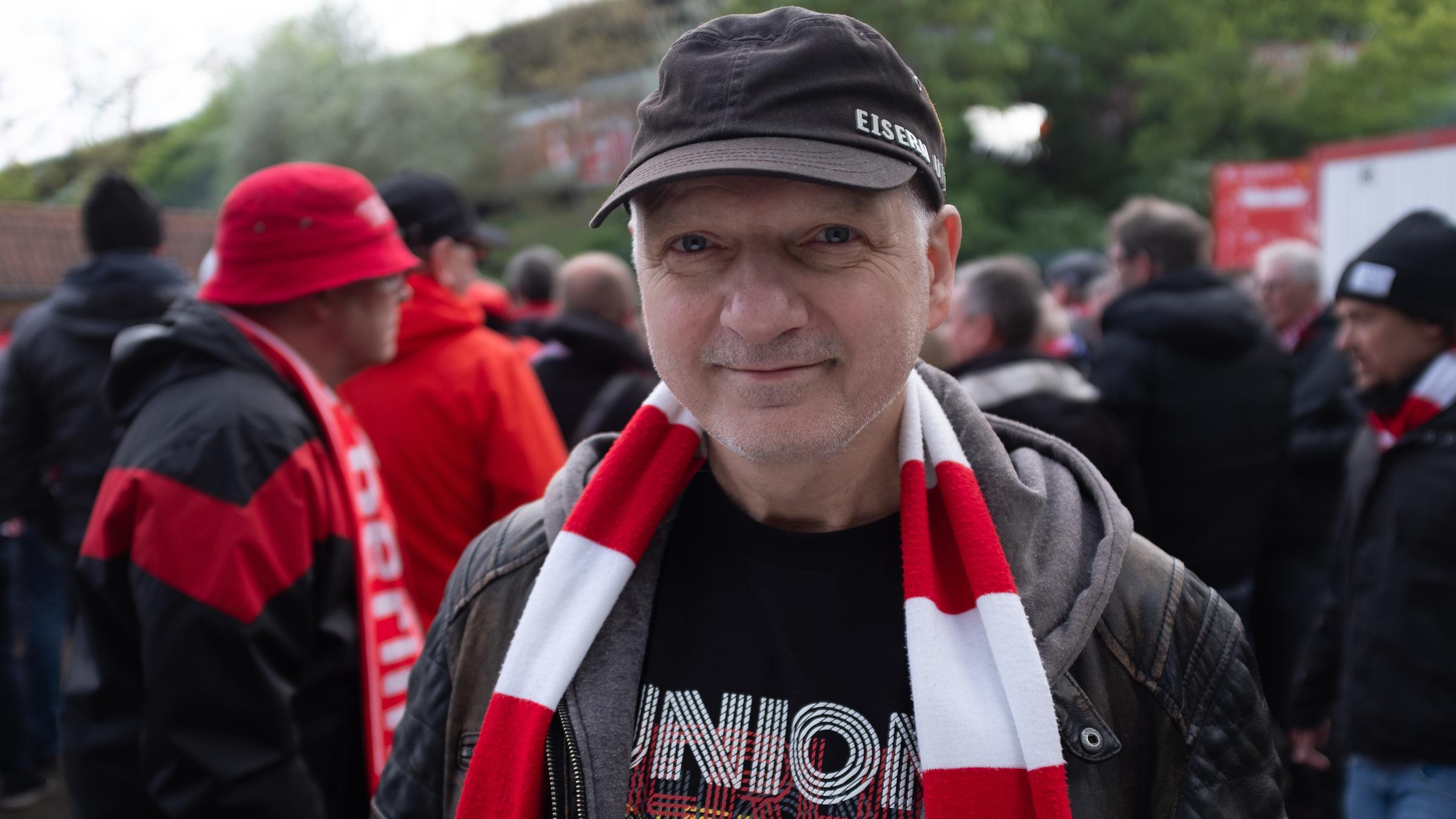 "Fußballhymnen. Schalalalala?": Mann mit Basecap und rot-weißem Fan-Schal vor einer Menschenmenge weiterer Fans.
