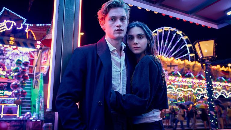 "Love Sucks" Drehstart. Ben (Damian Hardung) Zelda (Havana Braun) umarmen sich. Im Hintergrund ist eein Rummelplatz in bunter, nächtlicher Beleuchtung zu sehen.