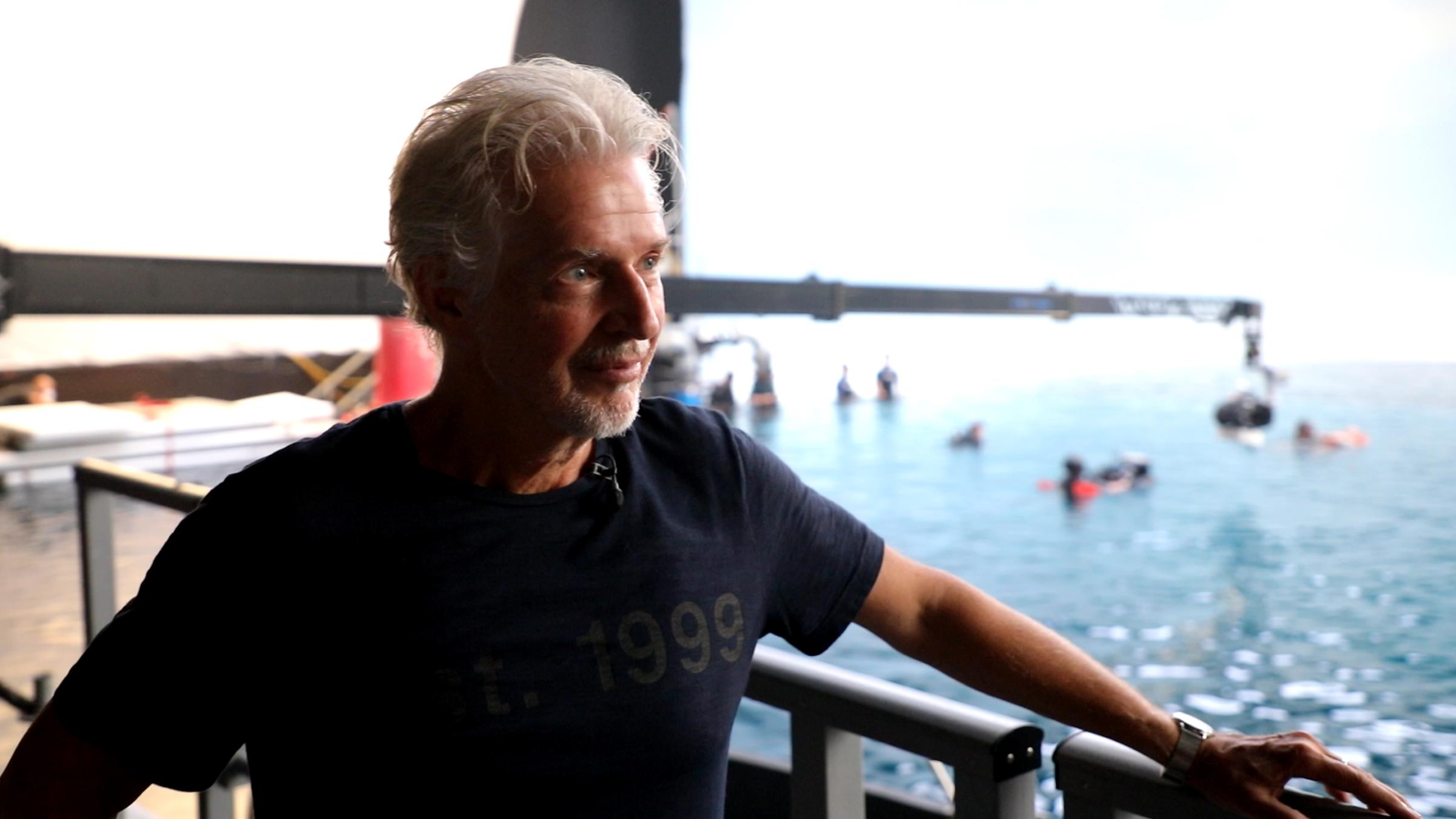 "Frank Schätzing - Mein Schwarm": Frank Schätzing steht an ein Geländer gelehnt und blickt zur Seite. Im Hintergrund ist ein Gewässer mit verschiedenen unscharfen Objekten im Wasser erkennbar, darüber ein Kamerakran.