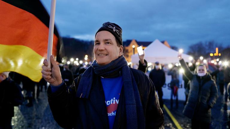 "Eine deutsche Partei": Ein lachender Mann mit Wollmütze und einem blauen Sweatshirt mit AfD-Aufdruck steht auf einem Platz in einer Anti-Corona-Demonstration und schwenkt eine Fahne.