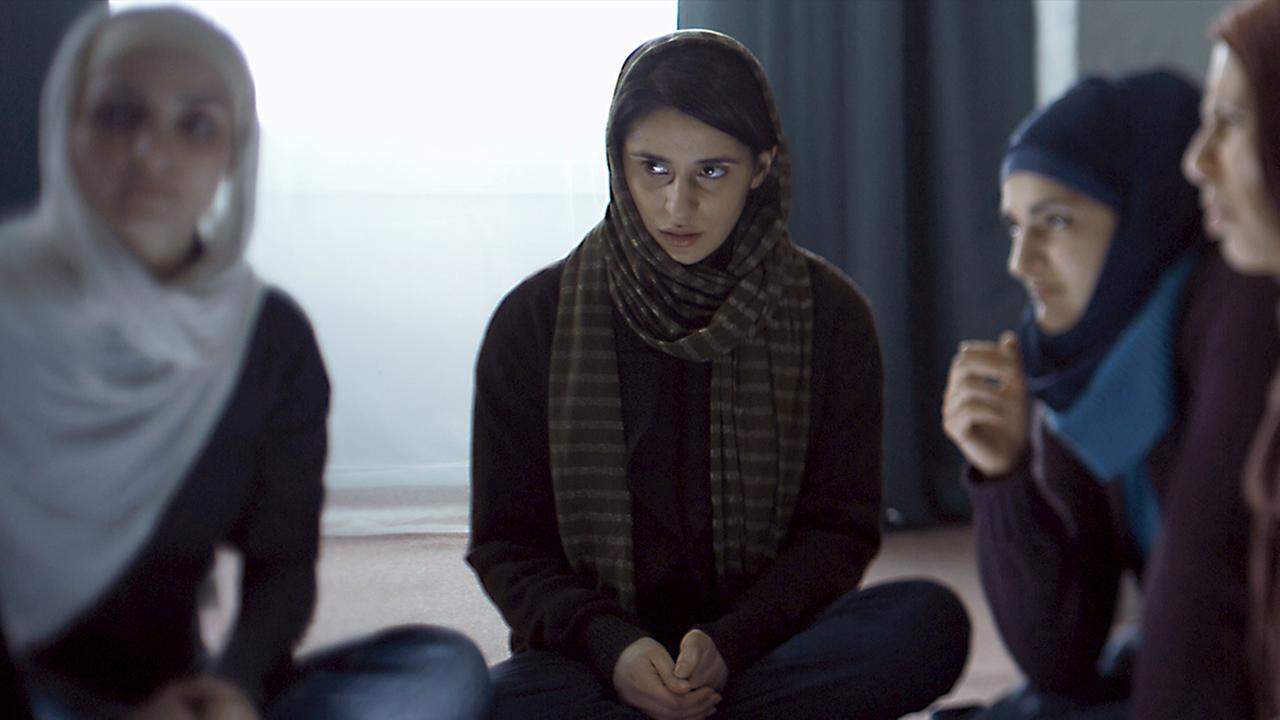 "Shahada": Maryam (Maryam Zaree), eine junge Frau mit dunklen Haaren und einem Tuch um den Kopf, sitzt mit anderen Frauen, die alle Kopftücher tragen, auf dem Boden. Sie schaut zu den Frauen, die sich unterhalten.