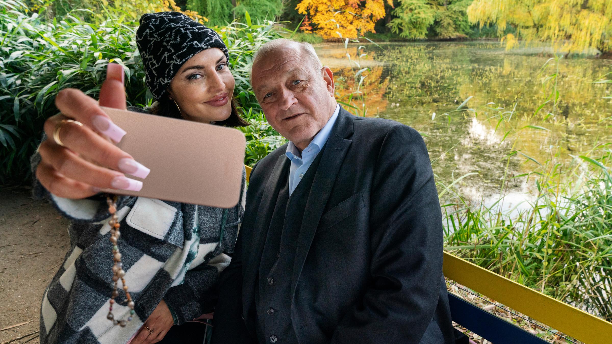 "Wilsberg – Folge mir": Wilsberg (Leonard Lansink) sitzt neben Bonnie von Laer (Johanna Ingelfinger) auf einer bunten Parkbank vor einem kleinen See. Bonnie holt ihr Handy vor sich und Wilsberg, um ein Selfie zu machen. Beide lächeln in die Handy-Kamera.