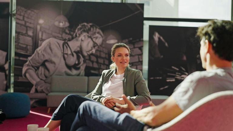 "Am Puls mit Sarah Tacke": Sarah Tacke sitzt mit einem Gesprächspartner vor einem Wandbild, auf dem eine arbeitende Frau in einer Fabrikhalle zu sehen ist.