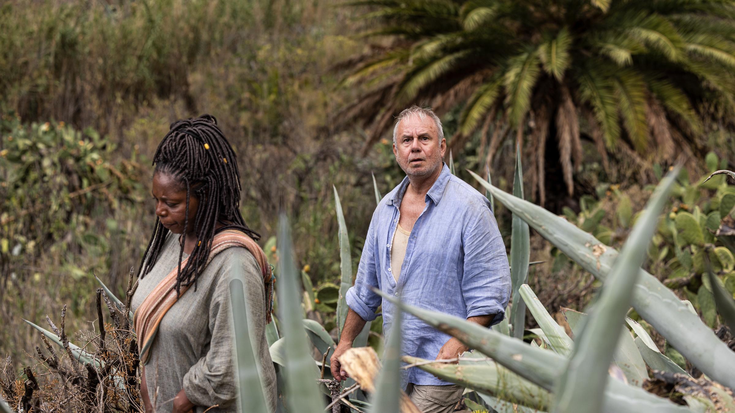 "Endlich Witwer - Über alle Berge": Hinter der Vegetation der Insel folgt Weiser (Joachim Król) erneut Soleil (Dela Dabulamanzi). Während ihr Blick nach unten geht, schaut Weiser beunruhigt und fragend auf.