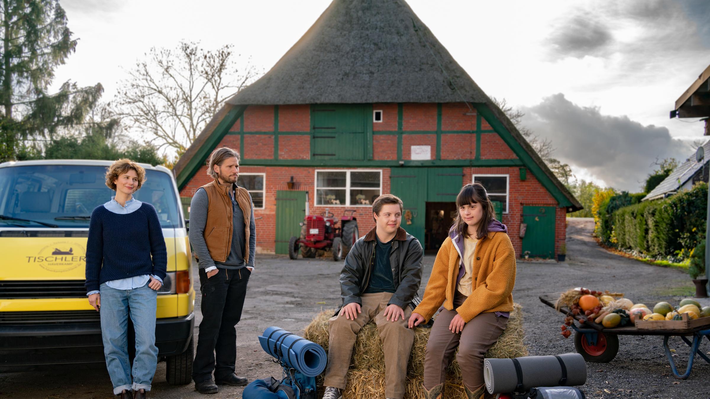 "Herzstolpern - Aufbruch nach Italien": Felix (Benjamin Raue) und Emma (Juliane Siebecke) sitzen auf einem großen Strohballen vor dem Mühlenhof, vor ihnen stehen jeweils ihre Rucksäcke mit Isomatten. Rechts hinter ihnen ist eine Schubkarre mit Kürbissen. Links daneben steht Elisa Jansen (Anna Maria Sturm) an Alex' Auto gelehnt. An ihrer Seite steht Alex Häverkamp (Sebastian Ströbel). Sie schauen beide lächelnd zu Felix und Emma.