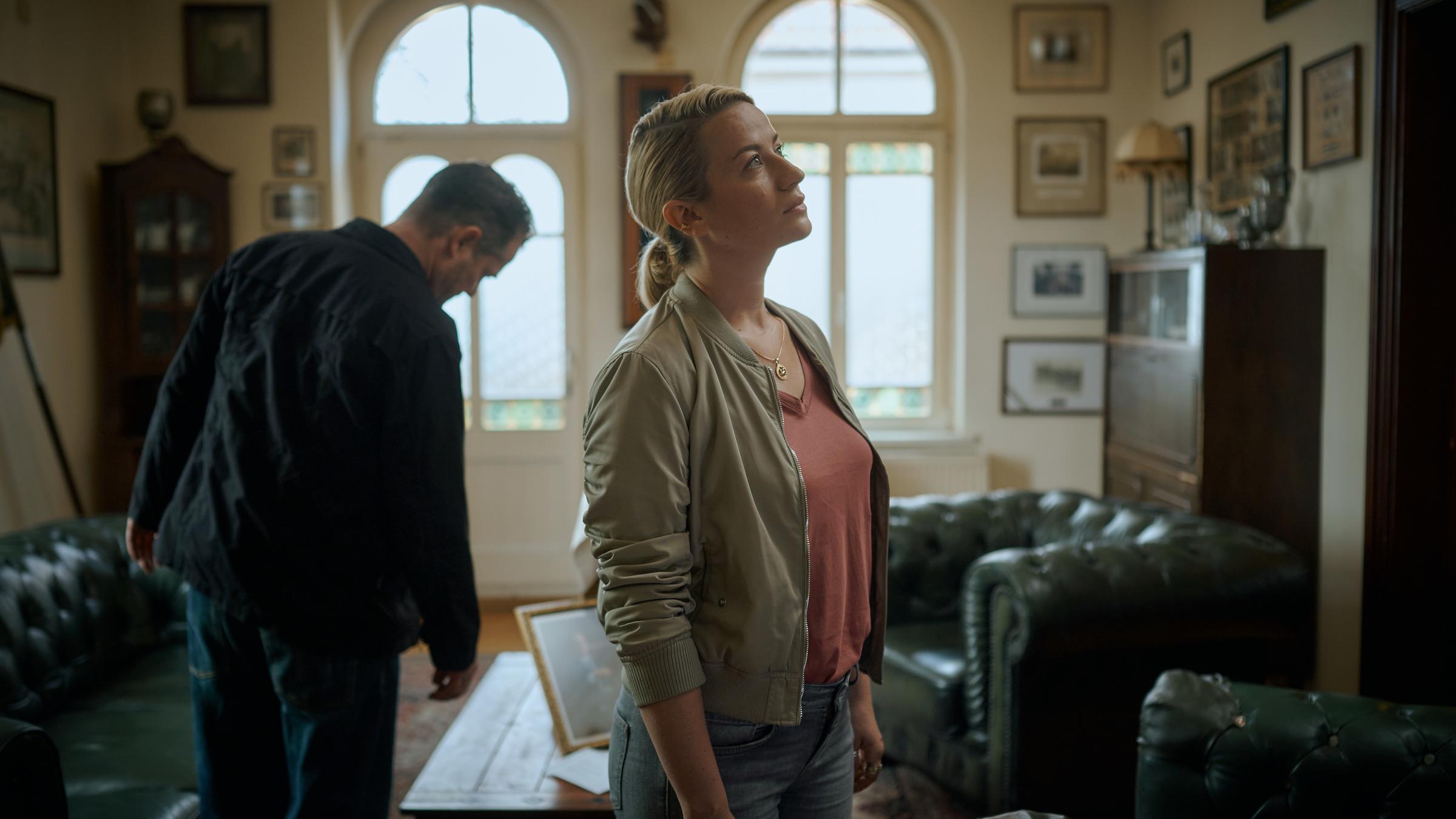 "Theresa Wolff - Der schönste Tag": Bruno Lewandowski (Aurel Manthei) und Theresa Wolff (Nina Gummich) sehen sich in einem altmodisch eingerichteten Zimmer um. An den Wänden hängen viele Bilderrahmen und in der Mitte des Raums steht ein kleiner Tisch mit zwei Sesseln. Lewandowski sieht sich einen Bilderrahmen auf dem Tisch an, während Wolff die Wand hochblickt.