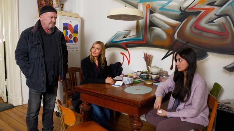 "Ein starkes Team - Kurierfahrt in den Tod": Linett (Stefanie Stappenbeck) und Aleyna (Sogol Faghani) sitzen an einem Küchentisch und blicken zu Otto (Florian Martens), der mit den Händen in den Jackentaschen neben einem freien Stuhl steht. An der Küchenwand im Hintergrund prangert ein großes Graffiti.