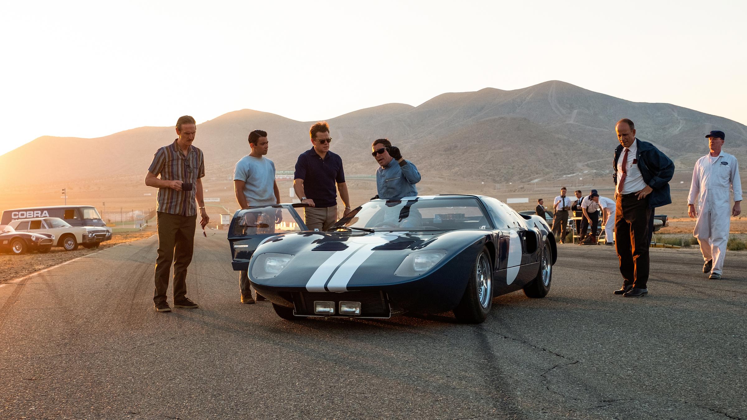 "Le Mans 66 - Gegen jede Chance": Mechaniker Phil (Ray McKinnon), Kollege Charlie (Jack McCullen), ihr Boss Carroll (Matt Damon) und Fahrer Ken (Christian Bale) stehen vor einem Rennwagen und sprechen miteinander. Weitere Personen sind rechts vom Wagen zu sehen. Im Hintergrund sieht man ein Landschaftspanorama und weitere Wagen an der Seite stehen.