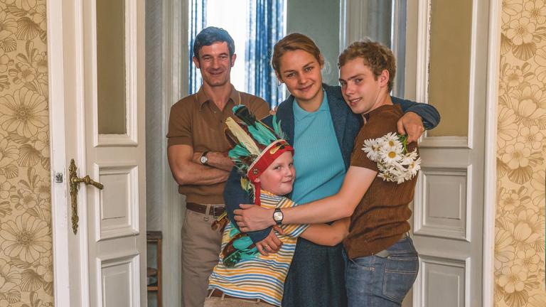 "Der Junge muss an die frische Luft": Mutter Margret (Luise Heyer), ihr Mann Heinz (Sönke Möhring), Hans-Peter (Julius Weckauf) und Matthes (Jan Lindner) stehen im Türrahmen zusammen und lächeln in die Kamera. Hans-Peter trägt einen gefiederten Häuptlingsschmuck auf dem Kopf.