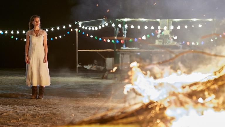 "Ostfriesenfeuer": Seitlich neben dem Osterfeuer steht Ann Kathrin Klaasen (Picco von Groote) in ihrem Brautkleid und schaut nachdenklich in die Flammen. Im Hintergrund sieht man mit Lichterketten beleuchtet, einen leer stehenden Getränkestand.