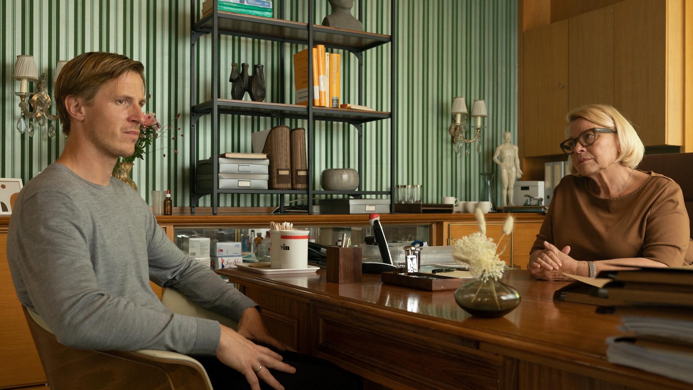 "Am Ende - Die Macht der Kränkung - David": David (Golo Euler) und Dr. Epstein (Margarethe Tiesel) sitzen um einen Schreibtisch versammelt und reden. Dr. Epstein sieht David an, dieser blickt in die Ferne. Auf dem Schreibtisch sind diverse Akten, eine Tasse und Deko zu sehen. Im Hintergrund sind ein Schrank und ein Sideboard zu sehen.