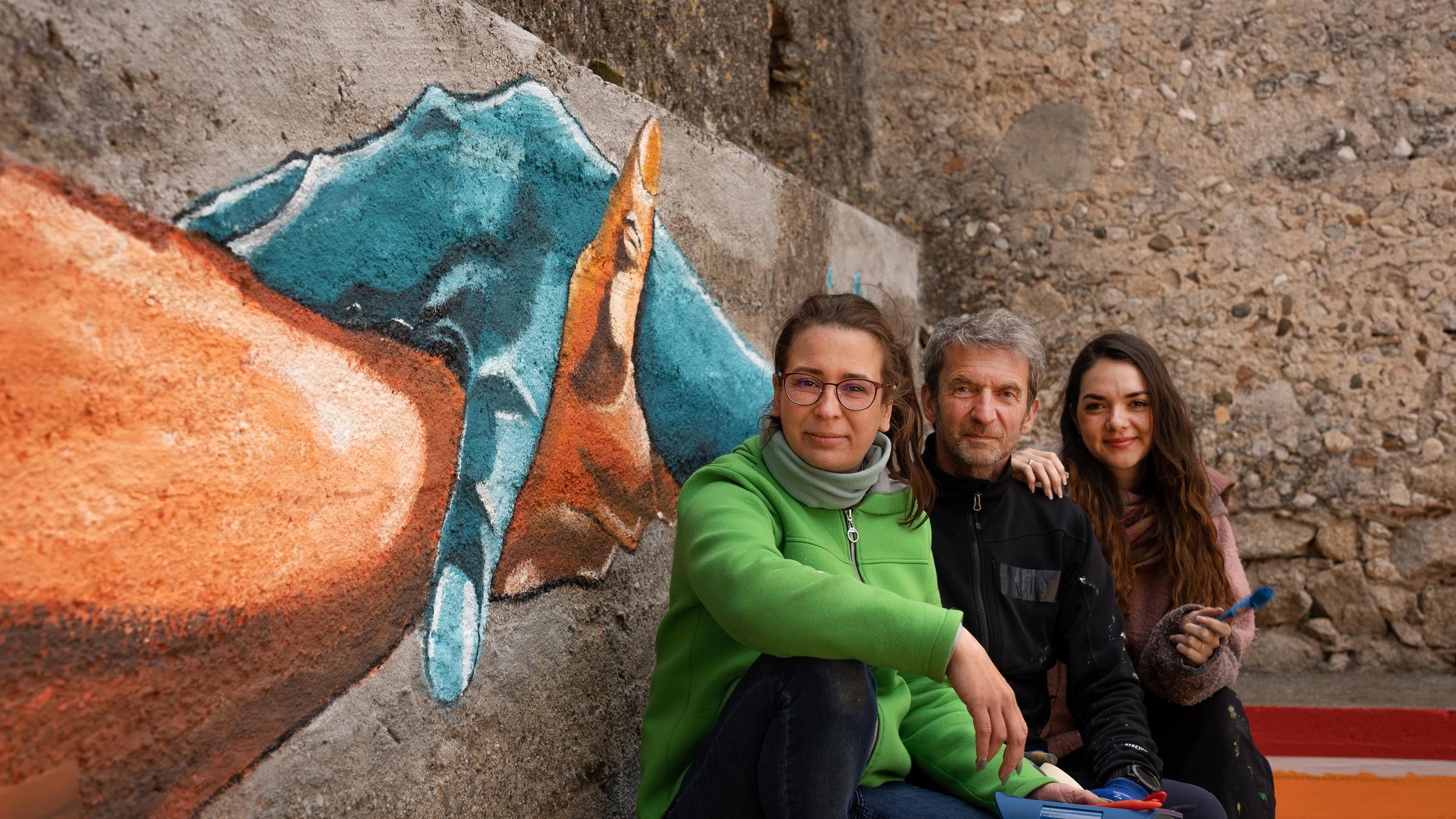 "Helfende Hände": Kristin, Gerhard und Philine blicken in die Kamera. Sie haben Malutensilien in den Händen. Sie sitzen vor einer Steinmauer mit zwei gemalten, sich haltenden Händen.