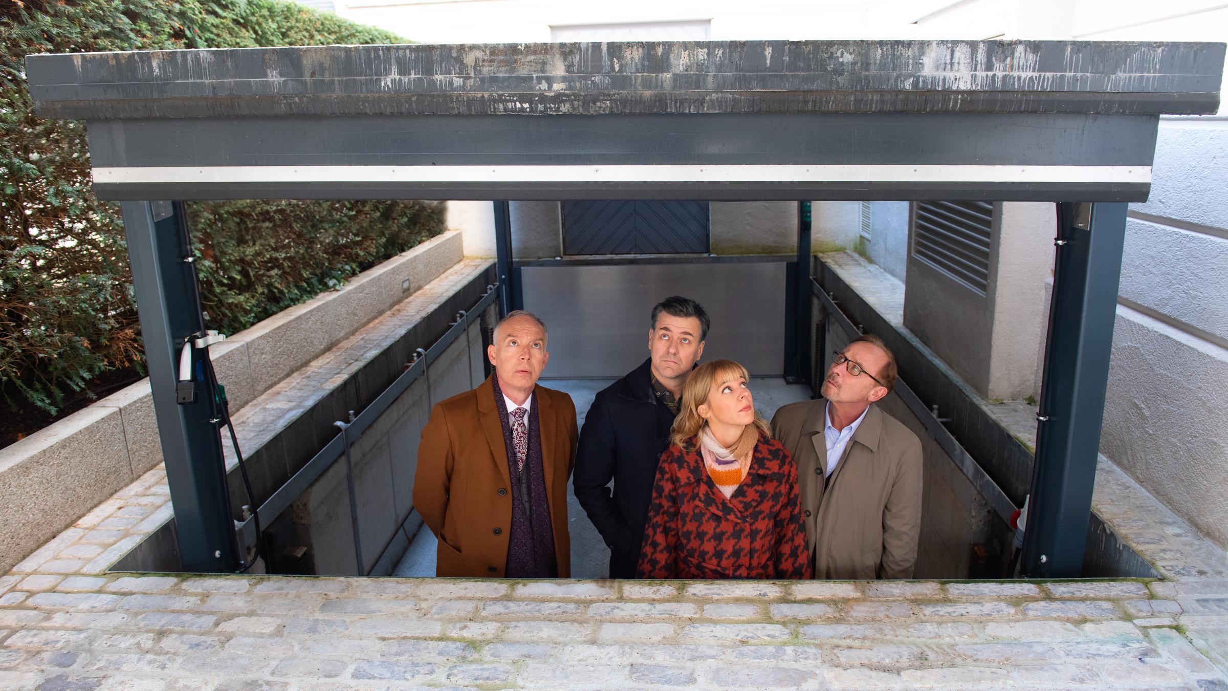 "München Mord - Der gute Mann vom Herzogpark": Helmut Zangel (Christoph Süß), Harald Neuhauser (Marcus Mittermeier), Angelika Flierl (Bernadette Heerwagen) und Ludwig Schaller (Alexander Held) blicken aus einem abgesenkten und überdachten Bereich nach oben. Sie sind umgeben von den Wänden einer Villa, sowie von einer hohen Hecke.