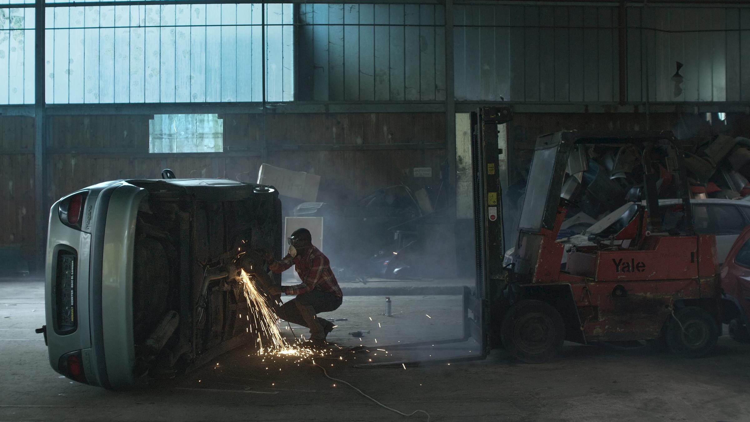 "Bewegungen eines nahen Bergs": In einer großen Montagehalle liegt ein graues Auto auf der Beifahrerseite, so dass der Mechaniker mit einem Schweiß- oder Flexgerät am Unterboden arbeiten kann.