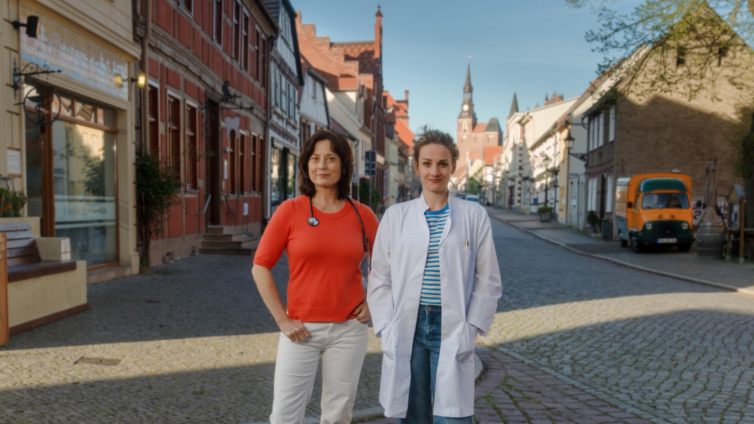 "Mit Herz und Holly - Diagnose Neustart": Neben der Arztpraxis stehen Dr. Katrin Herz (Inka Friedrich) und Dr. Holly Sass (Karoline Teska) auf der Straße. Katrin hat ein Stethoskop um den Hals gelegt, Holly trägt einen weißen Arztkittel. Beide blicken lächelnd in die Kamera.
