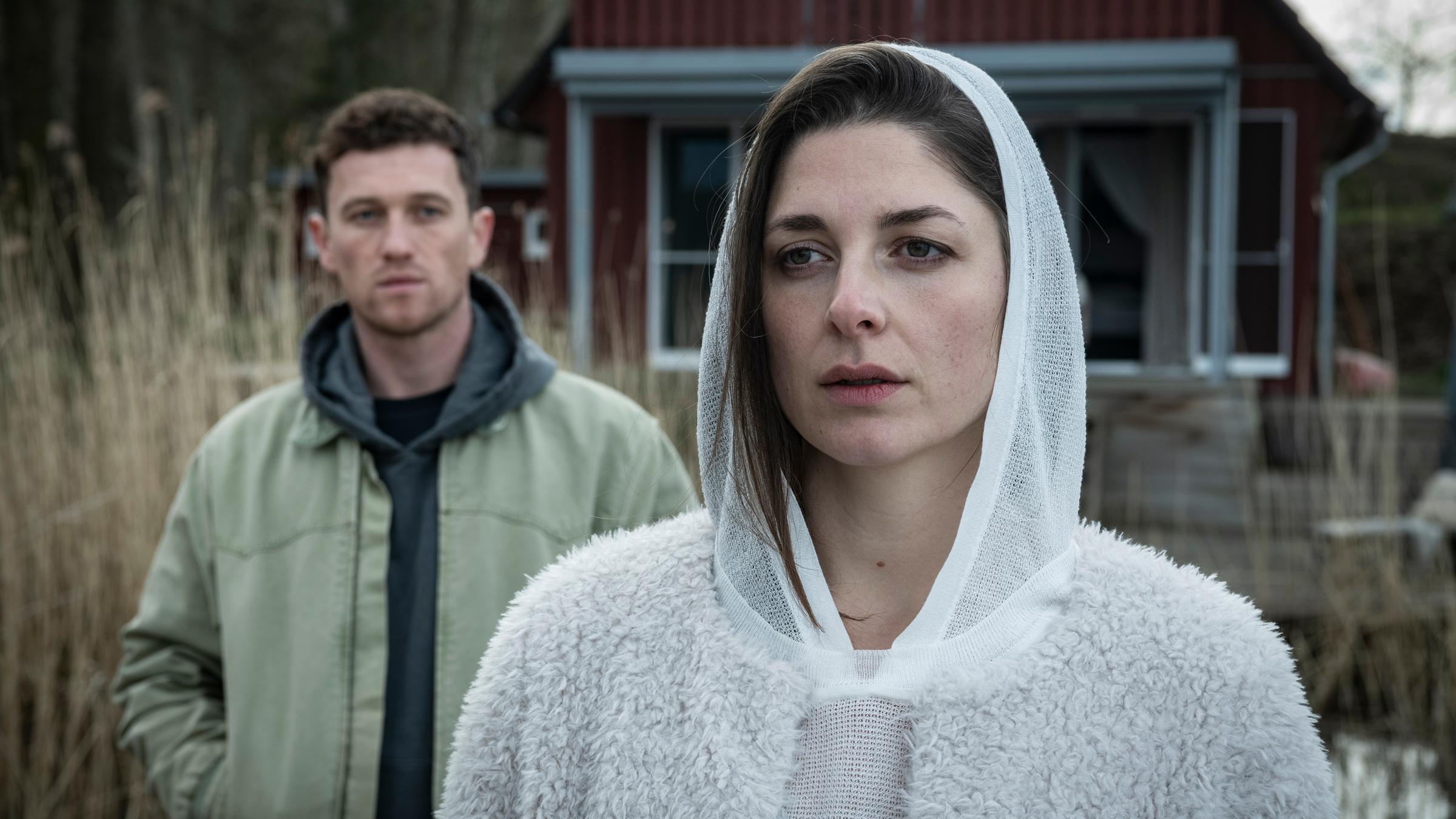 "Die Whistleblowerin": Galina Gromowa (Katharina Nesytowa) steht vor einem schwedischen Ferienhaus und schaut in die Ferne. Hinter ihr steht Tom Arndt (Artjom Gilz) und betrachtet sie.