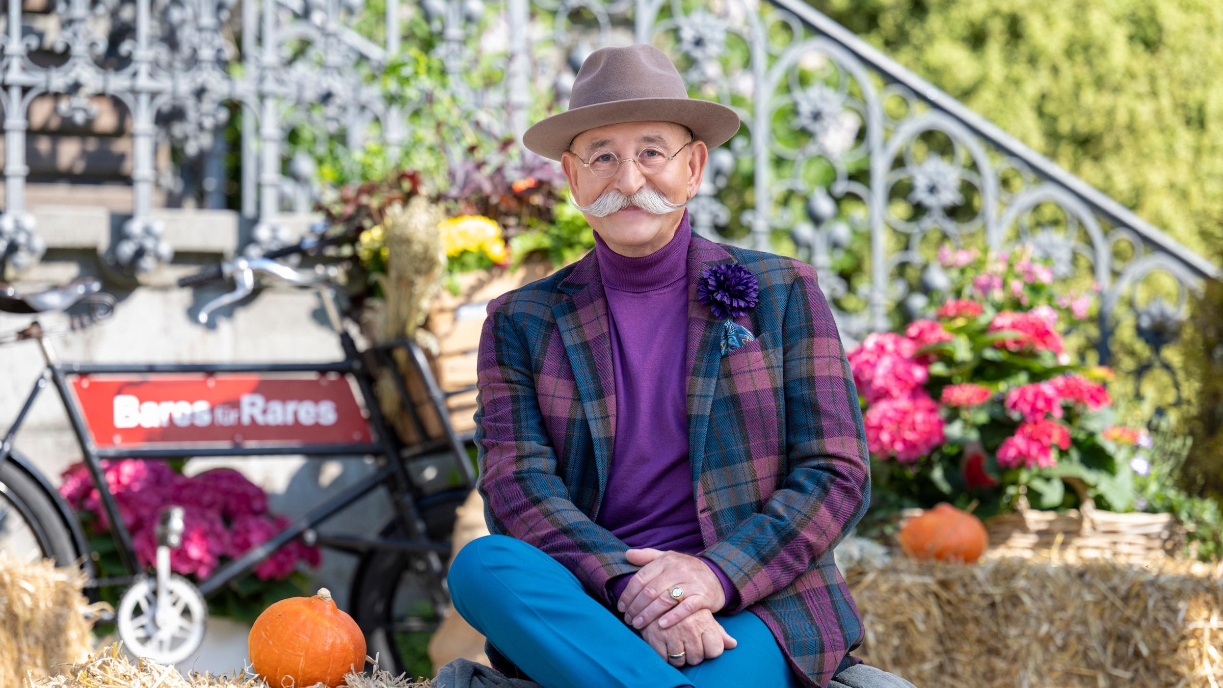 "Bares für Rares - Deutschlands größte Trödelshow": Horst Lichter sitzt auf einem Strohballen, schlägt die Beine übereinander und lächelt in die Kamera. Um ihn herum gibt es herbstliche Dekorationen vor einem schmiedeeisernem Treppengeländer. Im Hintergrund ist das Sendungslogo auf einem alten Fahrrad angebracht.
